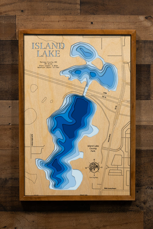 A meticulously detailed, laser-cut, multi-layered wooden topographical map of Island Lake in Ramsey County, Minnesota, showcasing the shallow 60-acre urban lake bisected by Interstate 694 with depth contours from the 5-foot average to the 12-foot maximum. The natural wood top layer frames approximately three miles of shoreline with Island Lake County Park facilities, the fishing pier, boat launch, and winter aeration system, while graduated blue layers create a gentle visual depth gradient across this belov