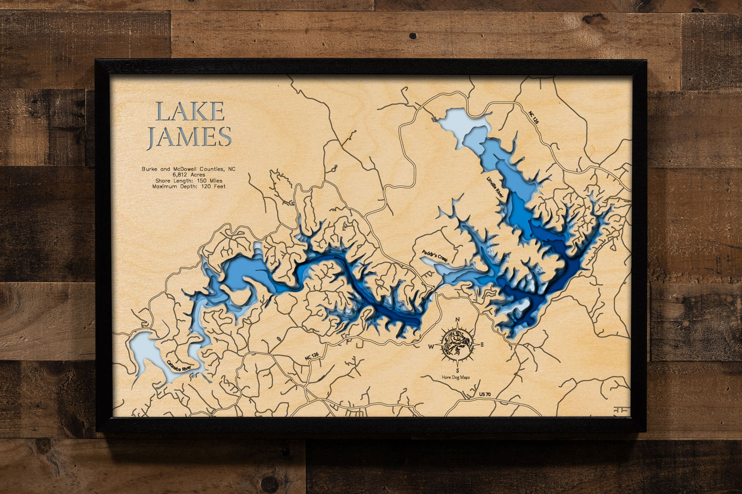 A laser-cut, layered wooden map of Lake James in North Carolina. The distinct shape of this large reservoir, with its numerous arms and 150-mile shoreline, is clearly visible as it spans Burke and McDowell Counties. The varying depths of the lake, reaching a maximum of 120 feet, are represented by different layers of wood, illustrating its impressive underwater topography.