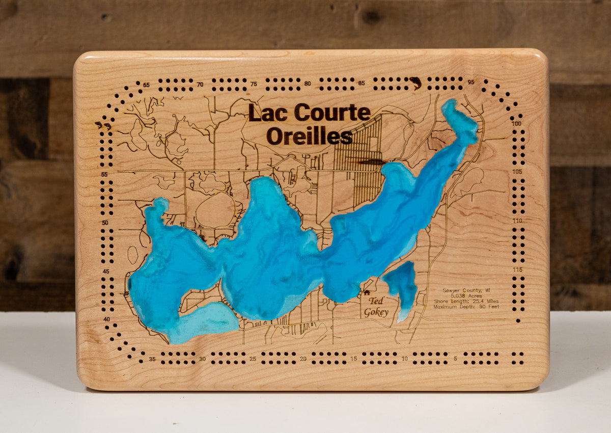 A cribbage board featuring an engraved map of Lac Courte Oreilles with bright blue resin-filled recesses, photographed against a rustic wood background.