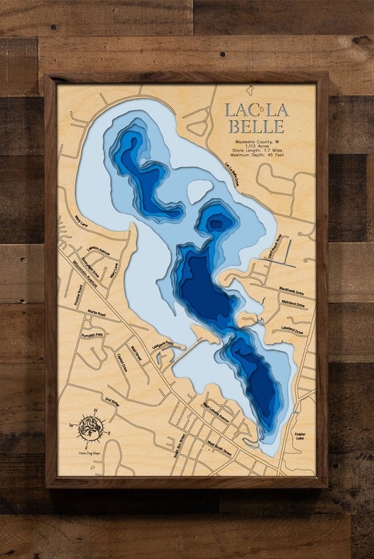 A laser-cut, layered wooden map of Lac La Belle in Waukesha County, Wisconsin. The distinct shape of this natural lake, with its 7.7-mile shoreline, is clearly visible within the landscape of southeastern Wisconsin. The varying depths of the lake, reaching a maximum of 45 feet, are represented by different layers of wood, illustrating its impressive underwater topography.