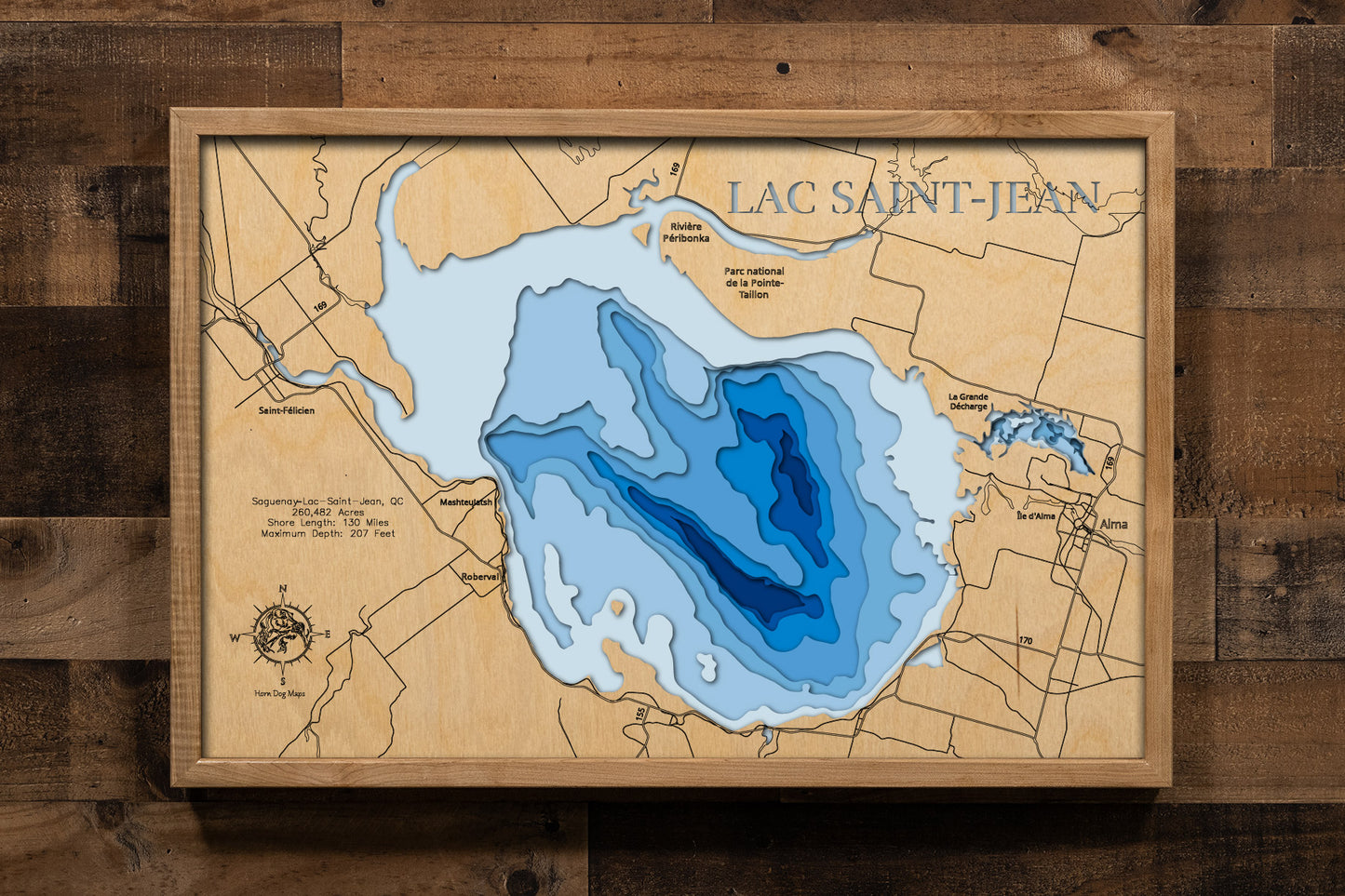 A magnificently detailed, laser-cut, multi-layered wooden topographical map of Lac Saint-Jean in Saguenay–Lac-Saint-Jean, Quebec, showcasing the expansive 260,482-acre inland sea measuring 45 by 34 kilometers with 130 miles of shoreline featuring picturesque villages and 42 kilometers of sandy beaches. The natural wood top layer frames the mouths of major rivers and the Véloroute des Bleuets route, while graduated blue layers create a stunning visual gradient from shallow waters to the 207-foot maximum dept