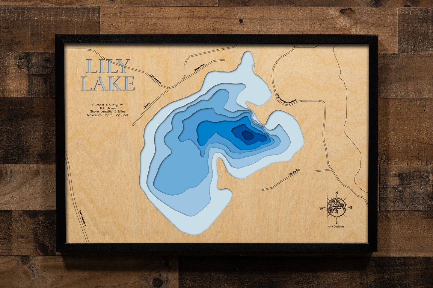 A meticulously detailed, laser-cut, multi-layered wooden topographical map of Lily Lake in Burnett County, Wisconsin, showcasing the 188-acre shallow lake with depth contours from the 5-foot average to the 22-foot maximum depth. The natural wood top layer frames approximately three miles of shoreline near Webster with the public boat landing and Lilly Lake Tavern location, while graduated blue layers create a gentle visual depth gradient across this clean, action-packed bluegill and bass fishing lake.