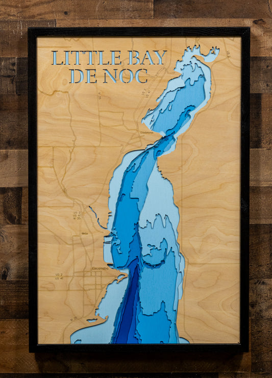 A framed wooden depth map of Little Bay De Noc, featuring light to dark blue water layers on a natural plywood background, mounted on a dark wood plank wall.