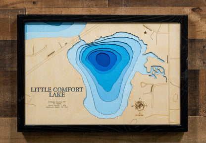 A framed wooden depth map of Little Comfort Lake, featuring layered blue water contours on a light wood background with a black frame, mounted on a rustic wood plank wall.