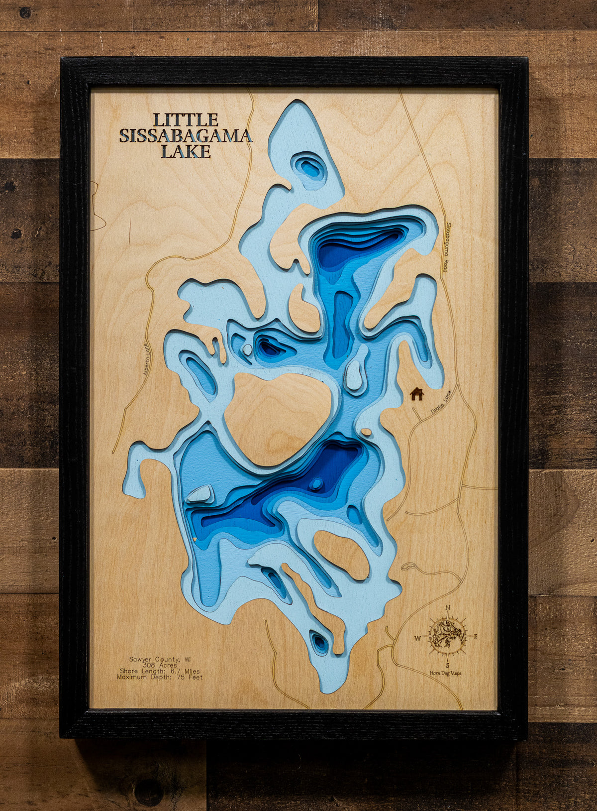 A framed wooden topographic map art piece of Little Sissabagama Lake, featuring layered blue and light blue resin in the recessed areas of a light wood background, enclosed in a black frame and mounted on a dark wood plank wall.