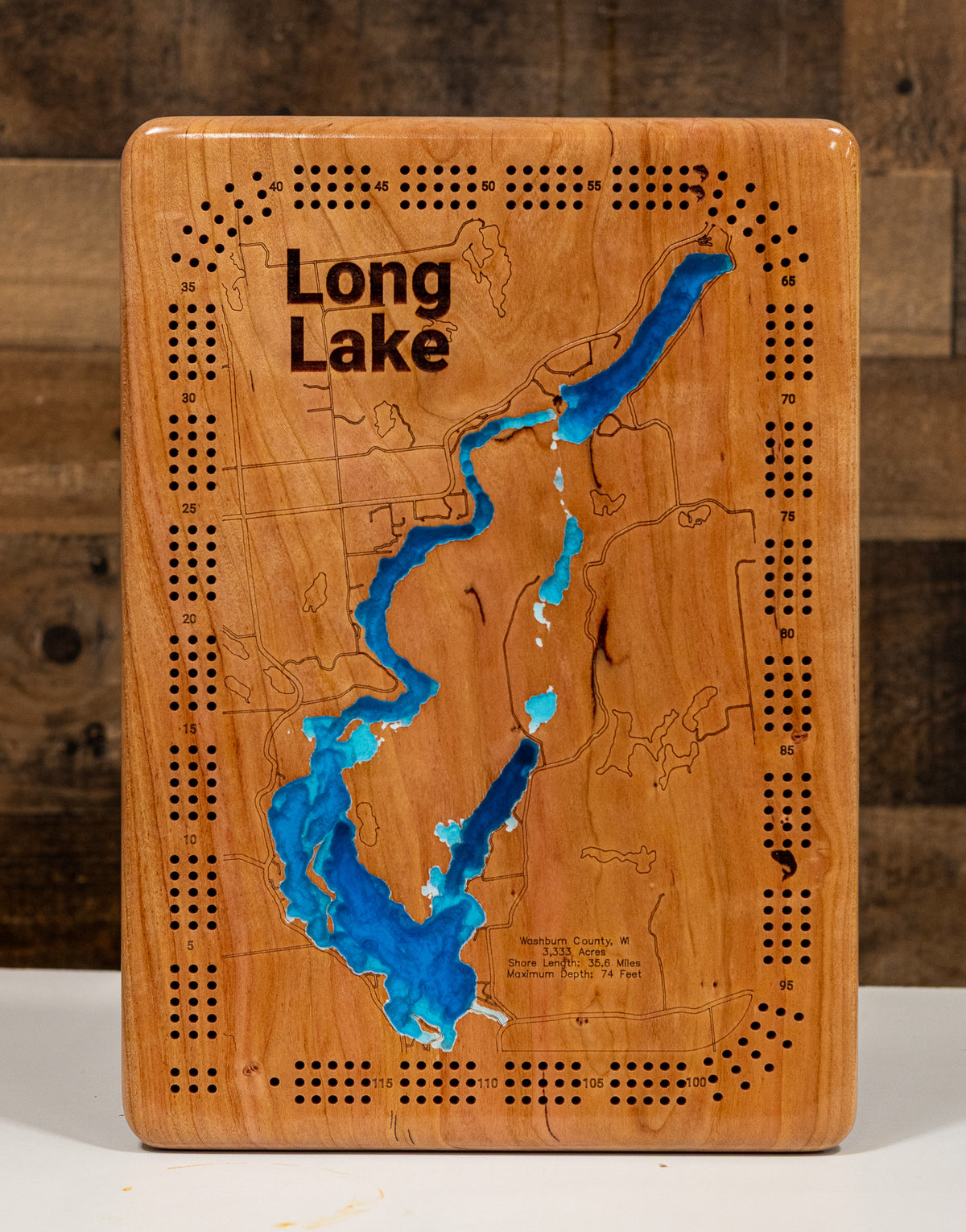 A  Long Lake cribbage board with laser-cut game pieces, featuring a natural wood grain finish and vibrant blue resin inlay of the lake, set against a rustic dark wood background.