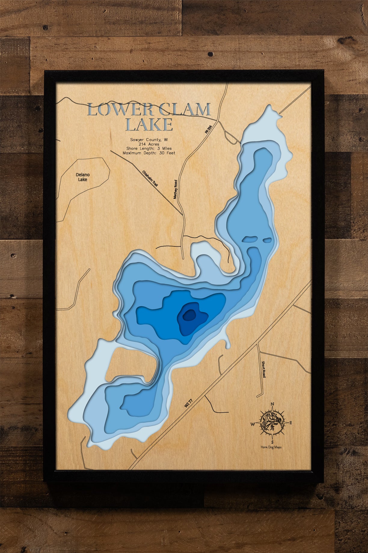 A meticulously detailed, laser-cut, multi-layered wooden topographical map of Lower Clam Lake in Sawyer County, Wisconsin, showcasing the 214-acre lake's irregular shape with multiple fishing bays and depth contours from sandy shallows to the 30-foot maximum depth. The natural wood top layer frames three miles of mostly undeveloped shoreline within the Chequamegon National Forest, while graduated blue layers create a striking visual depth gradient.