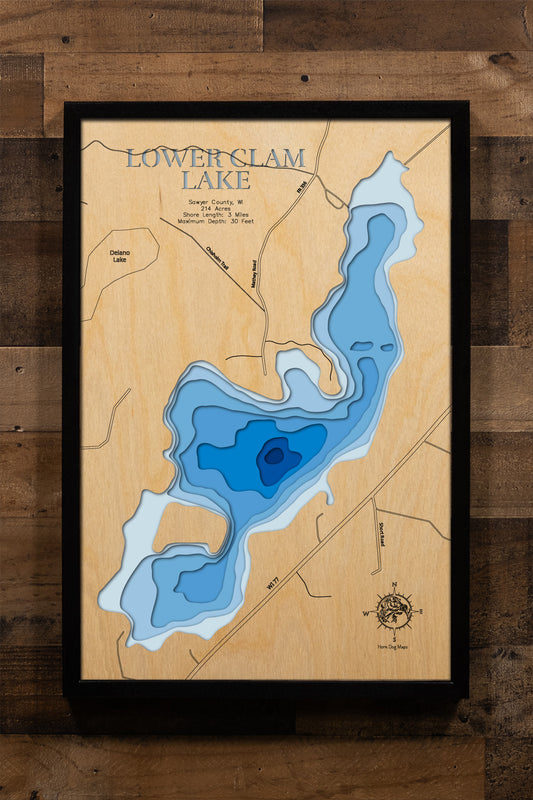 A meticulously detailed, laser-cut, multi-layered wooden topographical map of Lower Clam Lake in Sawyer County, Wisconsin, showcasing the 214-acre lake's irregular shape with multiple fishing bays and depth contours from sandy shallows to the 30-foot maximum depth. The natural wood top layer frames three miles of mostly undeveloped shoreline within the Chequamegon National Forest, while graduated blue layers create a striking visual depth gradient.