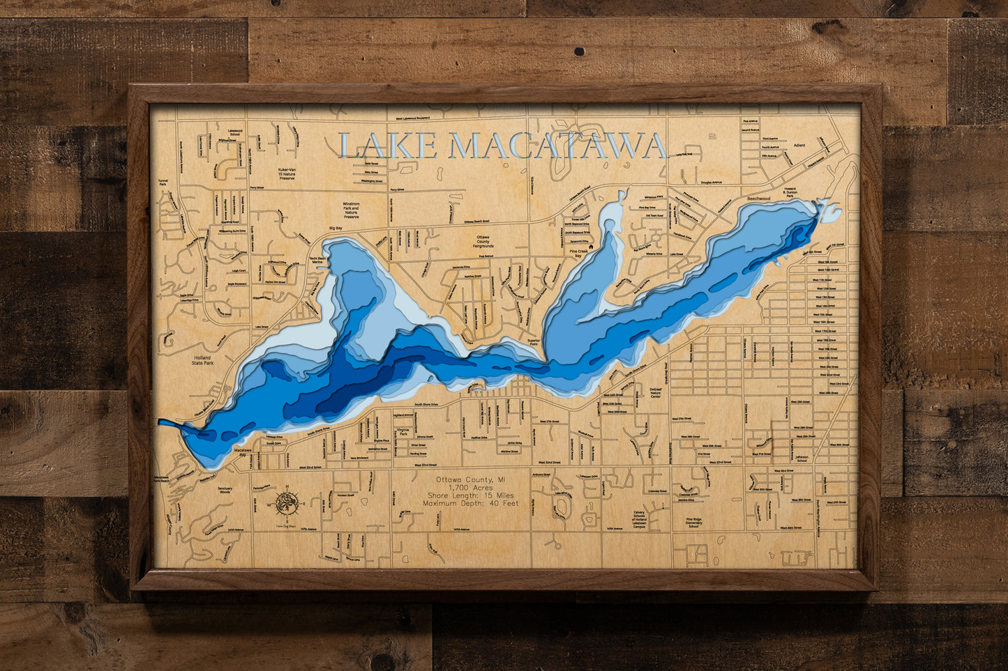 A beautifully detailed, laser-cut, multi-layered wooden topographical map of Lake Macatawa in Ottawa County, Michigan, showcasing the lake's 1,700-acre surface stretching six miles from the Macatawa River to Lake Michigan. The natural wood top layer frames 15 miles of shoreline including Big Bay, Pine Creek Bay, and Holland State Park, while graduated blue layers create a visual gradient from the 10-foot average depth to the 40-foot navigation channel.