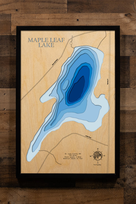 A meticulously crafted, laser-cut, multi-layered wooden map of Maple Leaf Lake in St. Louis County, Minnesota, featuring the lake's 100-acre surface area with precise depth contours showing the transition from shallow weedy bays to the 51-foot maximum depth. The natural wood top layer frames two miles of intricate shoreline detail, while graduated blue layers create a stunning depth gradient.