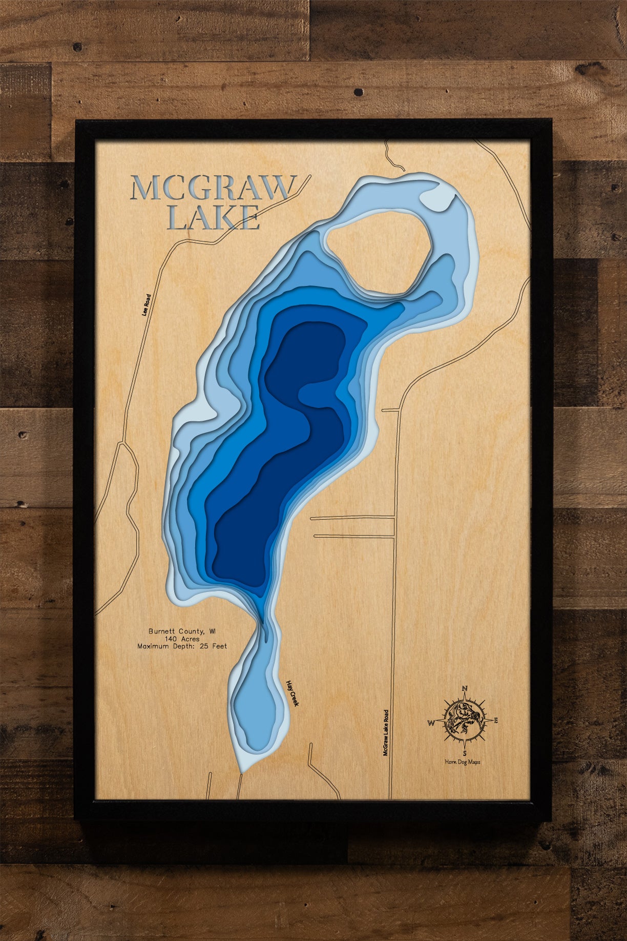 A precision laser-cut, multi-layered wooden map of McGraw Lake in Burnett County, Wisconsin. The natural wood top layer traces the lake's clean Northwoods shoreline with fine detail, while a light-to-dark blue gradient captures the lake's depth and dimension. A distinctive, rustic statement piece crafted for cabin walls, fishing lodges, or any Wisconsin lake-inspired living space.