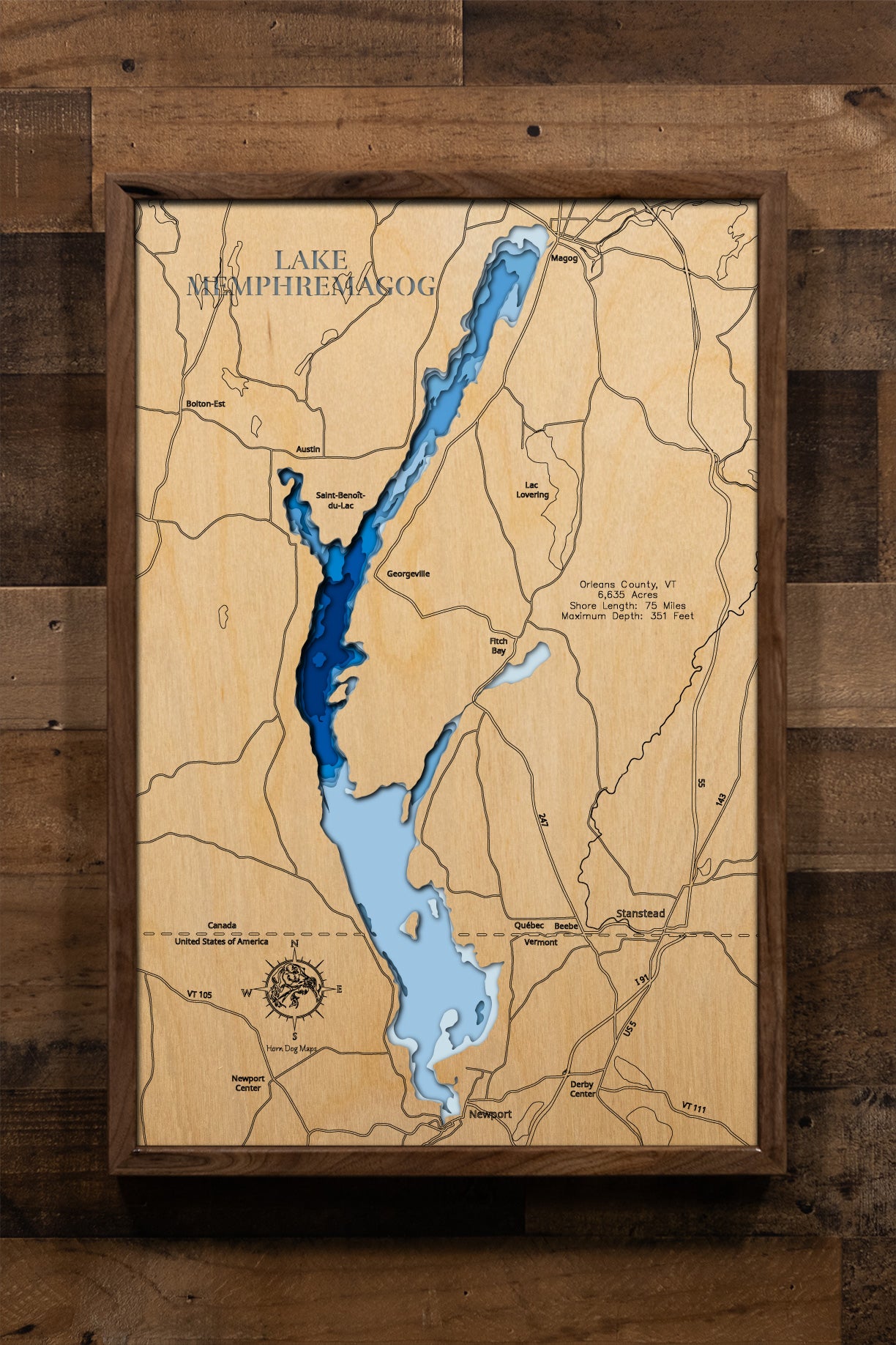 A finely detailed, laser-cut, multi-layered wooden topographic map of Lake Memphremagog spanning Orleans County, Vermont and Quebec, Canada. The intricate design captures the lake's distinctive 31-mile finger-like shape crossing the international border, 75 miles of scenic shoreline with numerous embayments including South Bay, Sargents Bay, and Fitch Bay, dramatic depth contours showing the shallow southern Vermont end contrasting with the 351-foot Canadian depths, and the 21 islands dotting the transbound