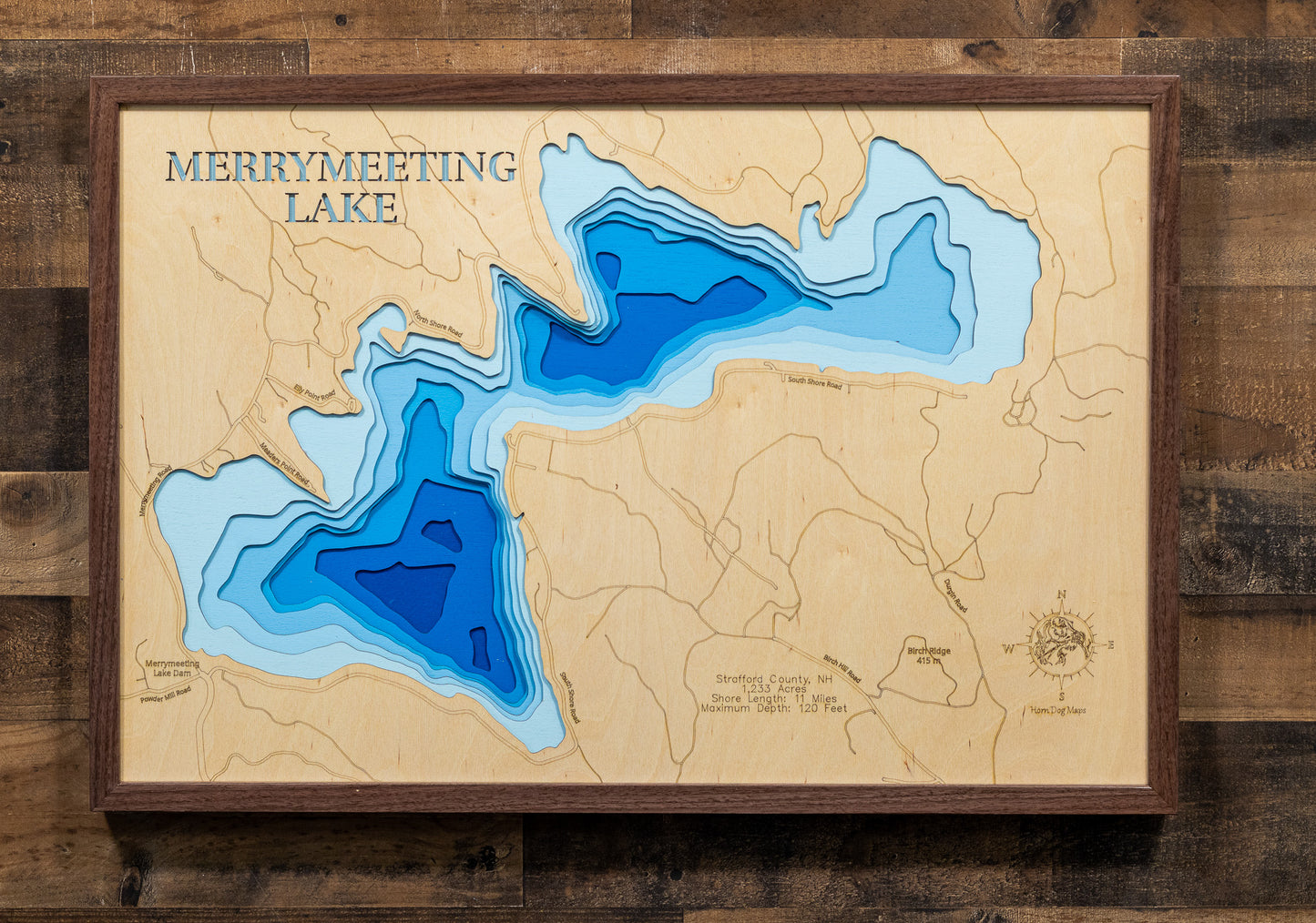 A magnificently detailed, laser-cut, multi-layered wooden topographical map of Merrymeeting Lake in Strafford County, New Hampshire, showcasing the 1,233-acre spring-fed lake's distinctive irregular hourglass shape with dramatic depth contours from the 29-foot average to the impressive 120-foot maximum depth. The natural wood top layer frames 11 miles of intricate shoreline including the dam, Merrymeeting River outlet to Lake Winnipesaukee, and protected conservation lands, while graduated blue layers creat