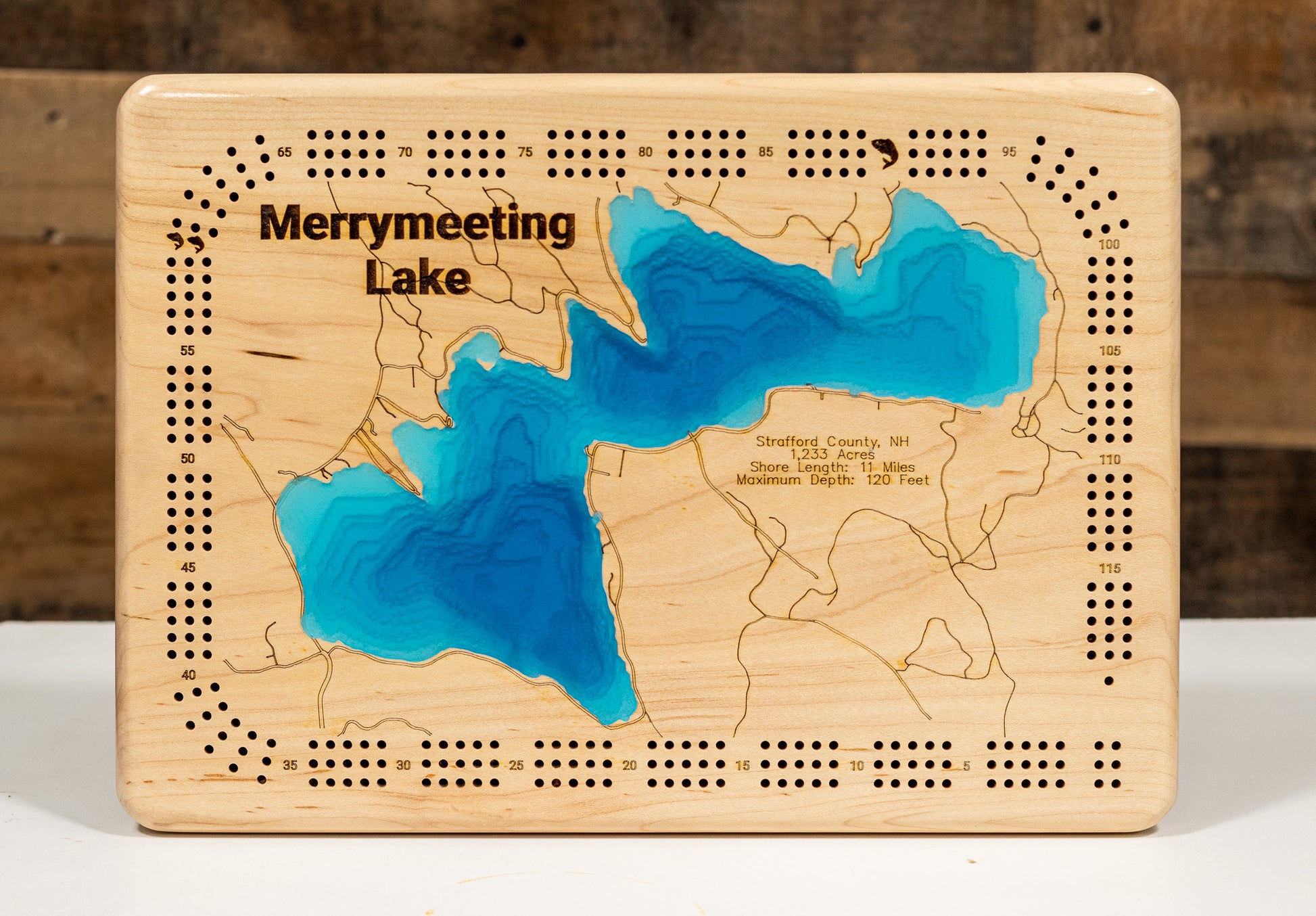 A magnificently detailed, laser-cut, multi-layered wooden topographical map of Merrymeeting Lake in Strafford County, New Hampshire, showcasing the 1,233-acre spring-fed lake's distinctive irregular hourglass shape with dramatic depth contours from the 29-foot average to the impressive 120-foot maximum depth. The natural wood top layer frames 11 miles of intricate shoreline including the dam, Merrymeeting River outlet to Lake Winnipesaukee, and protected conservation lands, while graduated blue layers creat