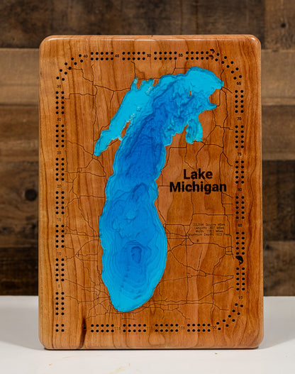 A square wooden board featuring an engraved map of Lake Michigan, with bright blue resin-filled contours and the words 'Lake Michigan' printed on the front. The board is photographed against a rustic dark wood background.