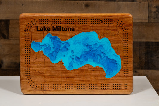 A cribbage board featuring a carved Lake Miltona map with bright blue resin inlay and black engraved text, displayed on a white surface against a rustic wood plank background.