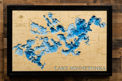 A framed 3D laser-etched map of Lake Minnetonka, Minnesota, featuring deep blue and light blue layered water depth contours on a beige background, enclosed in a black frame and mounted on a rustic wood wall.