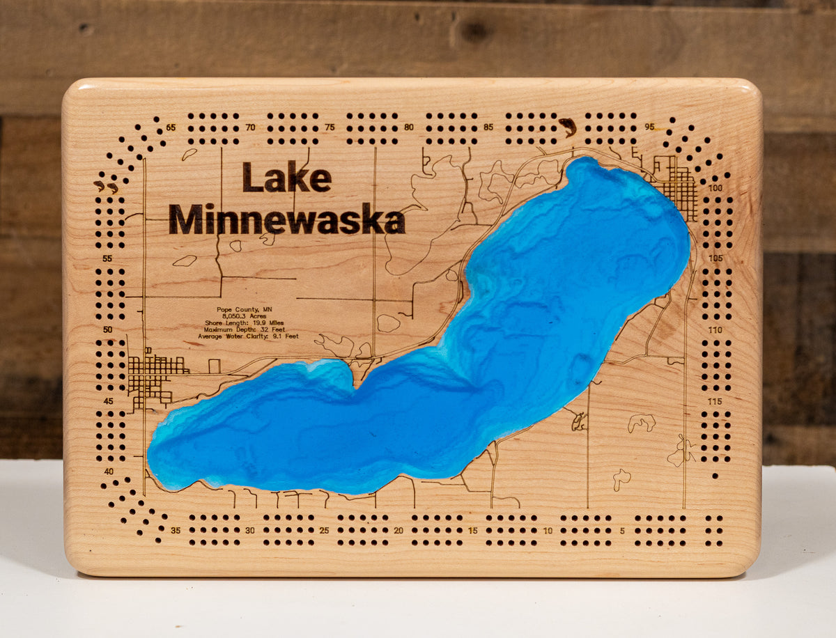 A cribbage board featuring an engraved map of Lake Minnewaska, with a carved blue resin lake shape and black text reading 'Lake Minnewaska,' shown on a maple surface against a dark wood background.