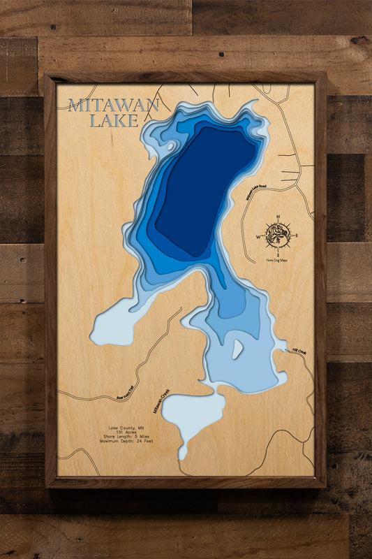 A meticulously detailed, laser-cut, multi-layered wooden topographical map of Mitawan Lake in Lake County, Minnesota, showcasing the 191-acre wilderness lake within Superior National Forest with depth contours from shallower areas to the 24-foot maximum depth. The natural wood top layer frames five miles of pristine shoreline accessible only by portage, while graduated blue layers create a gentle visual depth gradient across this remote gem.