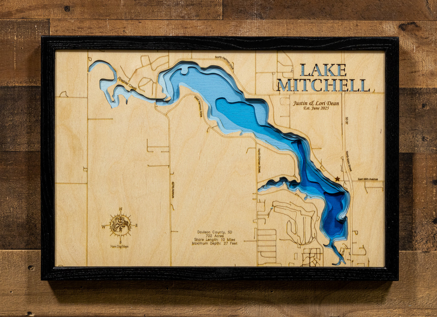 A carefully detailed laser-cut wooden topographic map of Lake Mitchell in Davison County, South Dakota, depicting the 702-acre man-made reservoir within Mitchell city limits, 10 miles of shoreline surrounded by 29 lakeside parks and access areas, depth contours showing the 27-foot maximum depth, and Firesteel Creek feeding from the west.