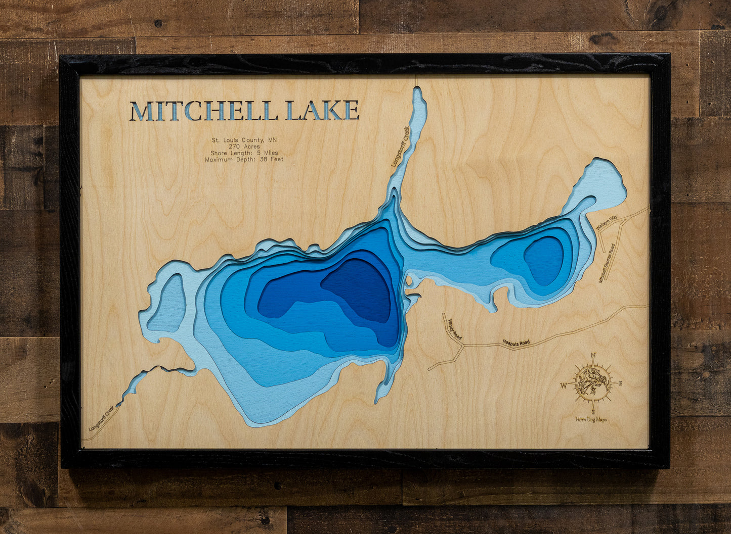A framed wooden Mitchell Lake depth map wall art piece, featuring layered blue wood sections representing water depth on a light wood background, mounted on a dark wood plank wall.