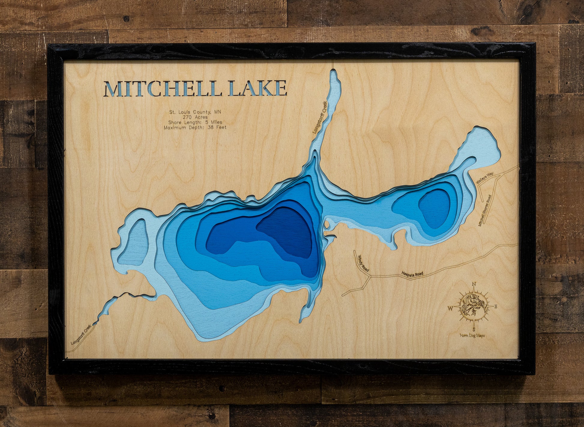 A framed wooden Mitchell Lake depth map wall art piece, featuring layered blue wood sections representing water depth on a light wood background, mounted on a dark wood plank wall.