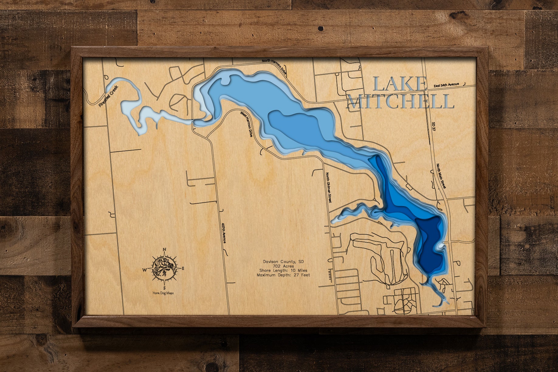 A carefully detailed laser-cut wooden topographic map of Lake Mitchell in Davison County, South Dakota, depicting the 702-acre man-made reservoir within Mitchell city limits, 10 miles of shoreline surrounded by 29 lakeside parks and access areas, depth contours showing the 27-foot maximum depth, and Firesteel Creek feeding from the west.