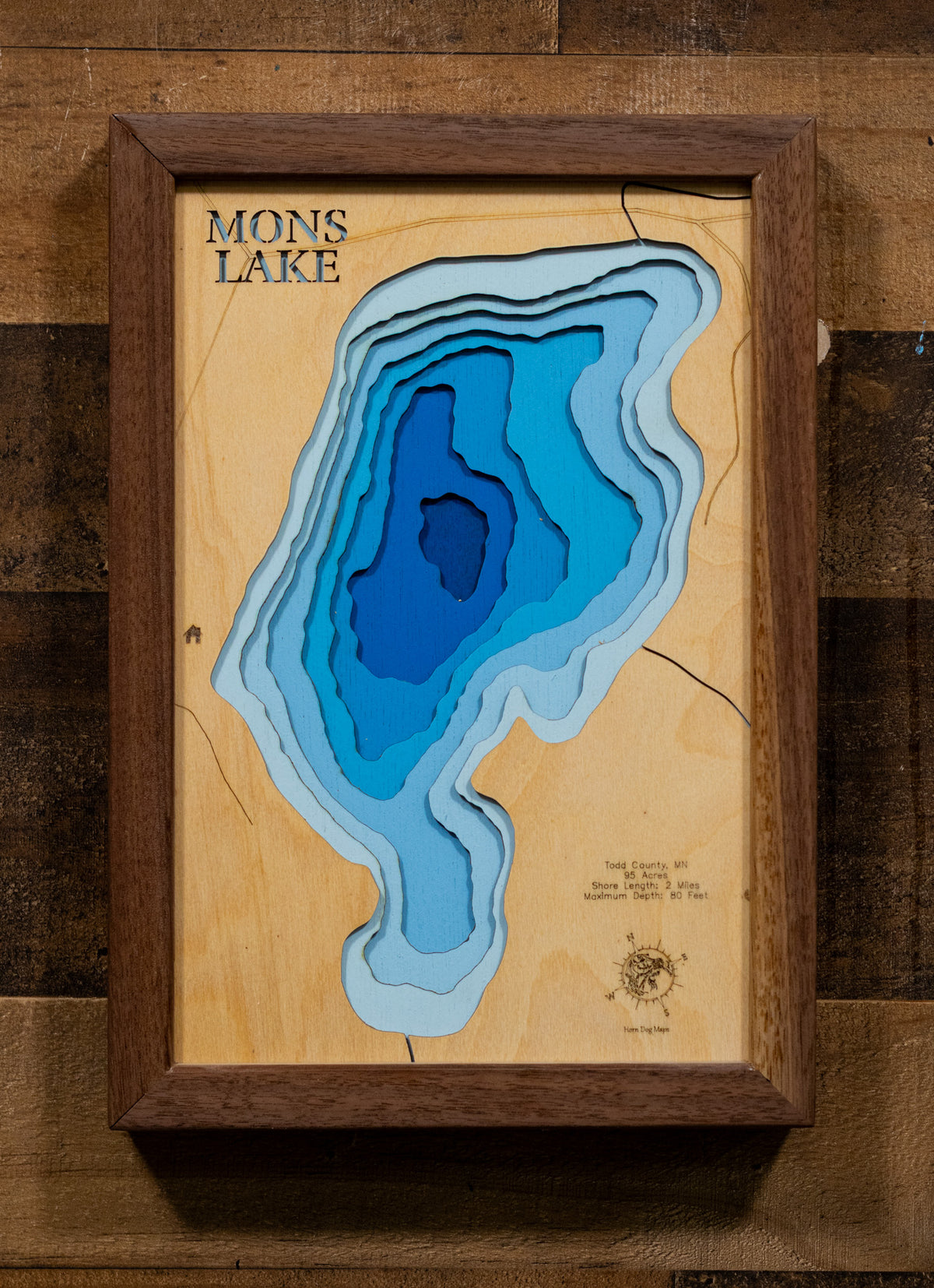 A precision laser-cut, multi-layered wooden map of Mons Lake in Todd County, Minnesota, a 95-acre lake near Long Prairie with a striking maximum depth of 80 feet. The natural wood top layer captures 2 miles of shoreline in fine detail, while a light-to-dark blue gradient beautifully conveys the lake's remarkable depth. A handcrafted statement piece for any Minnesota lake cabin, home, or outdoor enthusiast's wall.