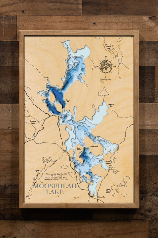 A laser-cut, layered wooden map of Moosehead Lake, located in Piscataquis County, Maine. The distinctive shape of Maine's largest lake is clearly visible within the northwestern Maine landscape. The varying depths of the lake, reaching a maximum of 246 feet, are represented by different layers of wood, illustrating its significant underwater topography.