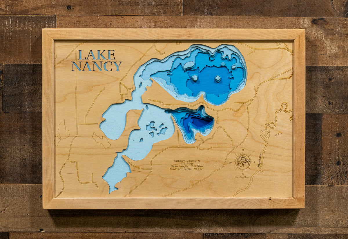 A 3D wooden map wall art piece of Lake Nancy, featuring layered light and dark blue wood cutouts on a natural wood background, mounted on a rustic dark wood plank wall.
