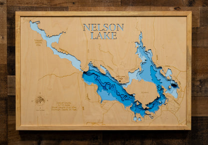 A 3D wooden topographic map of Nelson Lake, featuring layered blue and light blue wood sections to represent water depth, mounted on a rustic dark wood plank wall.