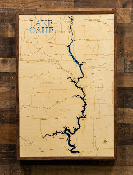 A framed wooden map of Lake Oahe, featuring a natural light wood background, detailed state gridlines, and the blue silhouette of the lake. The piece is housed in a dark brown wooden frame and mounted on a rustic dark wood plank wall.