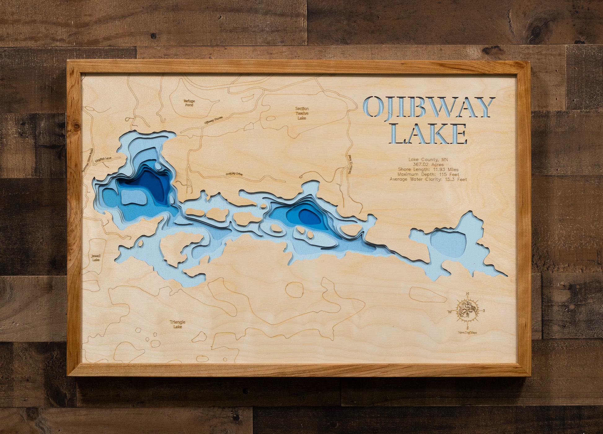 A laser-cut, layered wooden map of Ojibway Lake in Lake County, Minnesota. The distinct, irregular shape of this natural lake, with its 11.93-mile shoreline, is clearly visible within the forested landscape of northeastern Minnesota. The varying depths of the lake, reaching a maximum of 115 feet, are represented by different layers of wood, illustrating its impressive underwater topography.