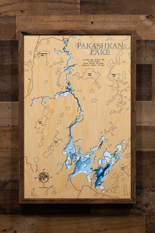 A magnificently detailed, laser-cut, multi-layered wooden topographical map of Pakashkan Lake in Thunder Bay District, Ontario, showcasing the sprawling 13,141-acre surface with three distinct basins and intricate 60 miles of wilderness shoreline. The natural wood top layer frames rocky reefs, protected bays, and the Grew-Weaver Canoe Route connections, while graduated blue layers create a stunning visual gradient from shallow river inlets to the 70-foot maximum depth.