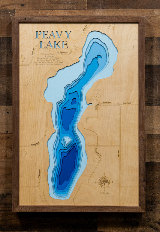 A framed wooden depth map of Peavy Lake, featuring layered blue and light blue concentric shapes representing water depth on a natural wood background, enclosed in a dark wood frame against a rustic dark wood wall.
