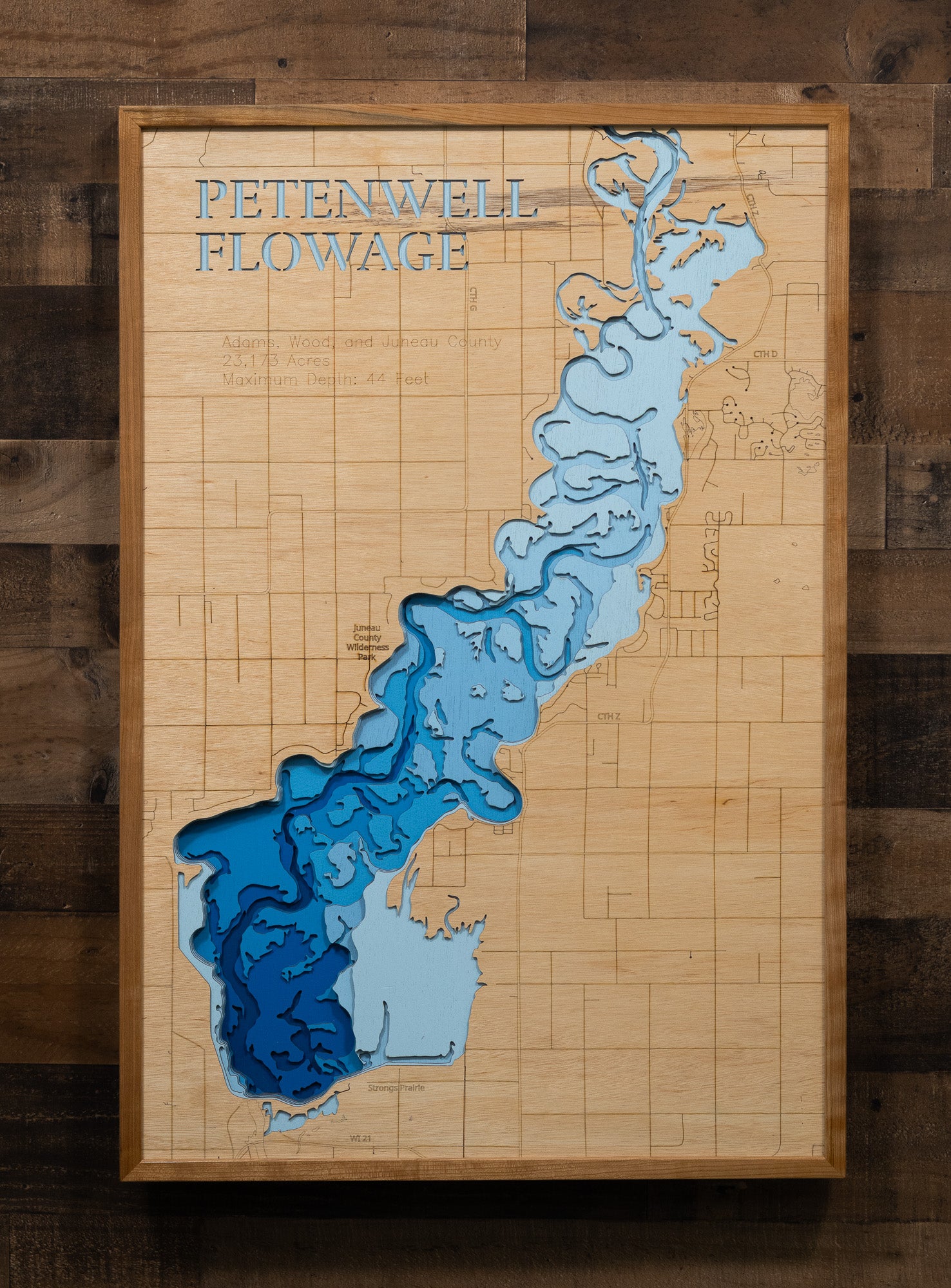 A laser-cut, layered wooden map of Petenwell Flowage in Adams, Juneau, and Wood counties, Wisconsin. The distinct shape of this vast artificial lake on the Wisconsin River, with its 101.07-mile shoreline, is clearly visible within the forested landscape of central Wisconsin. The varying depths of the flowage, reaching a maximum of 44 feet, are represented by different layers of wood, illustrating its underwater topography.