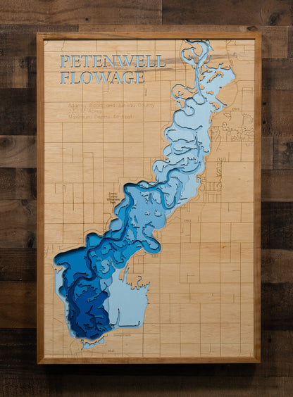 A laser-cut, layered wooden map of Petenwell Flowage in Adams, Juneau, and Wood counties, Wisconsin. The distinct shape of this vast artificial lake on the Wisconsin River, with its 101.07-mile shoreline, is clearly visible within the forested landscape of central Wisconsin. The varying depths of the flowage, reaching a maximum of 44 feet, are represented by different layers of wood, illustrating its underwater topography.
