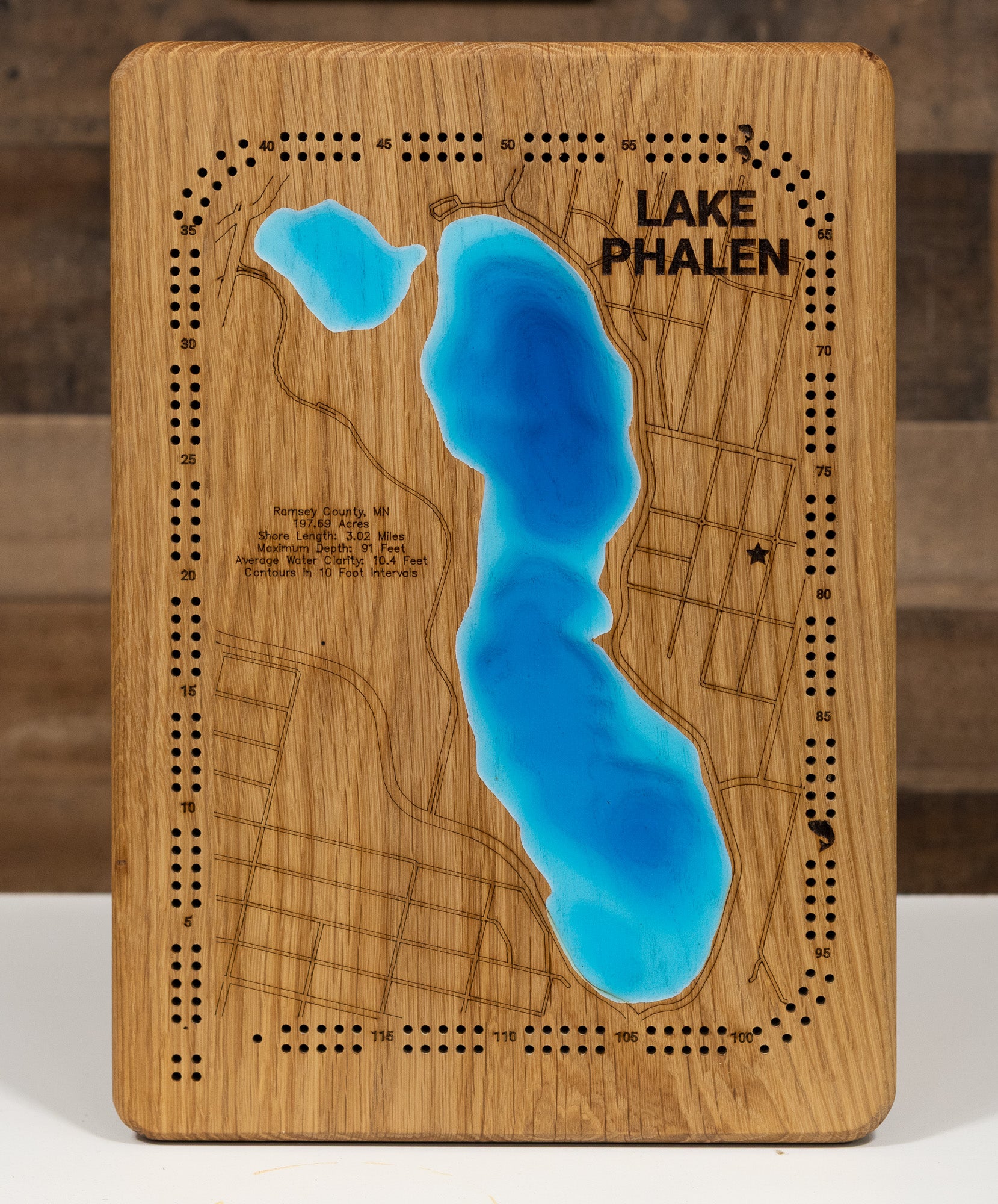 A laser-cut, layered wooden map of Lake Phalen in Ramsey County, Minnesota. The shape of the lake, with its 3.02-mile shoreline, is visible within the suburban landscape of Ramsey County. The varying depths of the lake, reaching a maximum of 91 feet and detailed by 10-foot contour intervals, are represented by different layers of wood, illustrating its underwater topography.