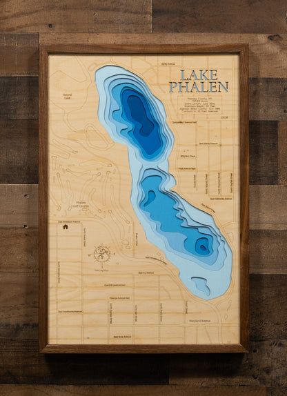 A laser-cut, layered wooden map of Lake Phalen in Ramsey County, Minnesota. The shape of the lake, with its 3.02-mile shoreline, is visible within the suburban landscape of Ramsey County. The varying depths of the lake, reaching a maximum of 91 feet and detailed by 10-foot contour intervals, are represented by different layers of wood, illustrating its underwater topography.
