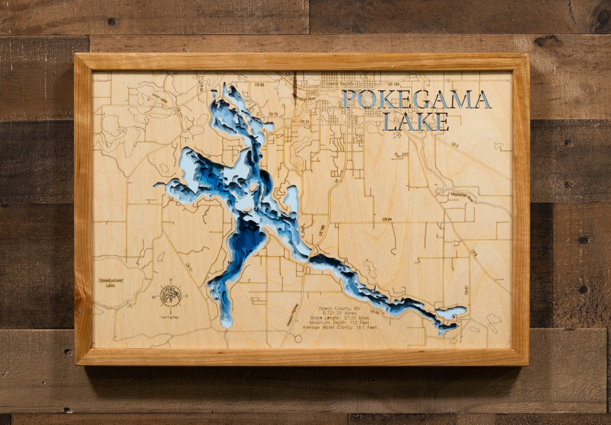 A laser-cut, layered wooden map of Pokegama Lake in Itasca County, Minnesota. The distinct, complex shape of this large natural lake, with its 58.26-mile shoreline, is clearly visible within the forested landscape of northern Minnesota. The varying depths of the lake, reaching a remarkable maximum of 110 feet, are represented by different layers of wood, illustrating its impressive underwater topography.