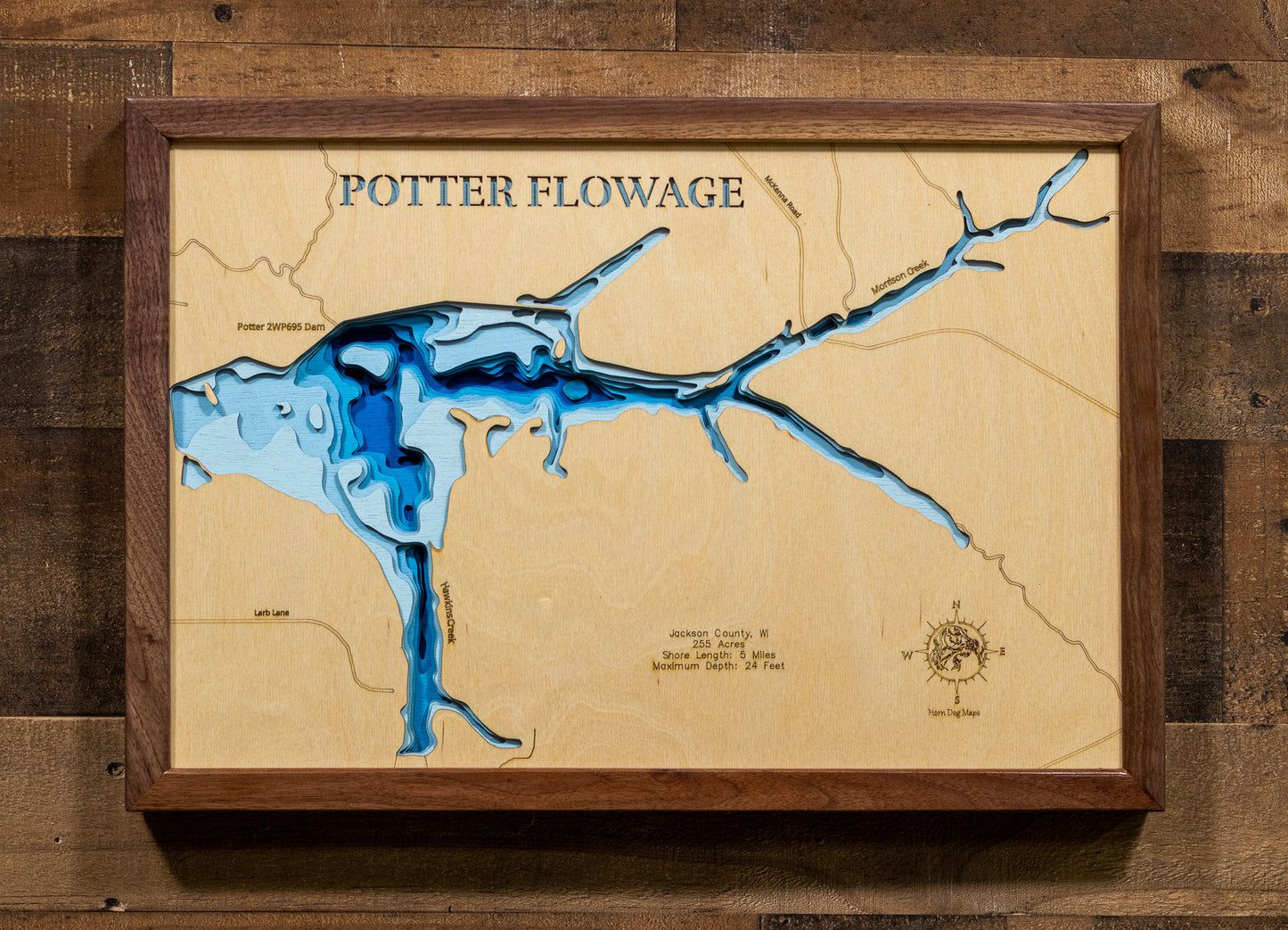 A meticulously detailed, laser-cut, multi-layered wooden topographical map of Potter Flowage in Jackson County, Wisconsin, showcasing the 255-acre impoundment's distinctive T-shape with McKenna and Morrison Creek inlets, numerous bays along approximately five miles of pine-lined shoreline, and extensive lily pad beds. The natural wood top layer frames the dark-water flowage with depth contours from the 7-foot average to the 24-foot maximum near the dam, while graduated blue layers create a gentle visual dep