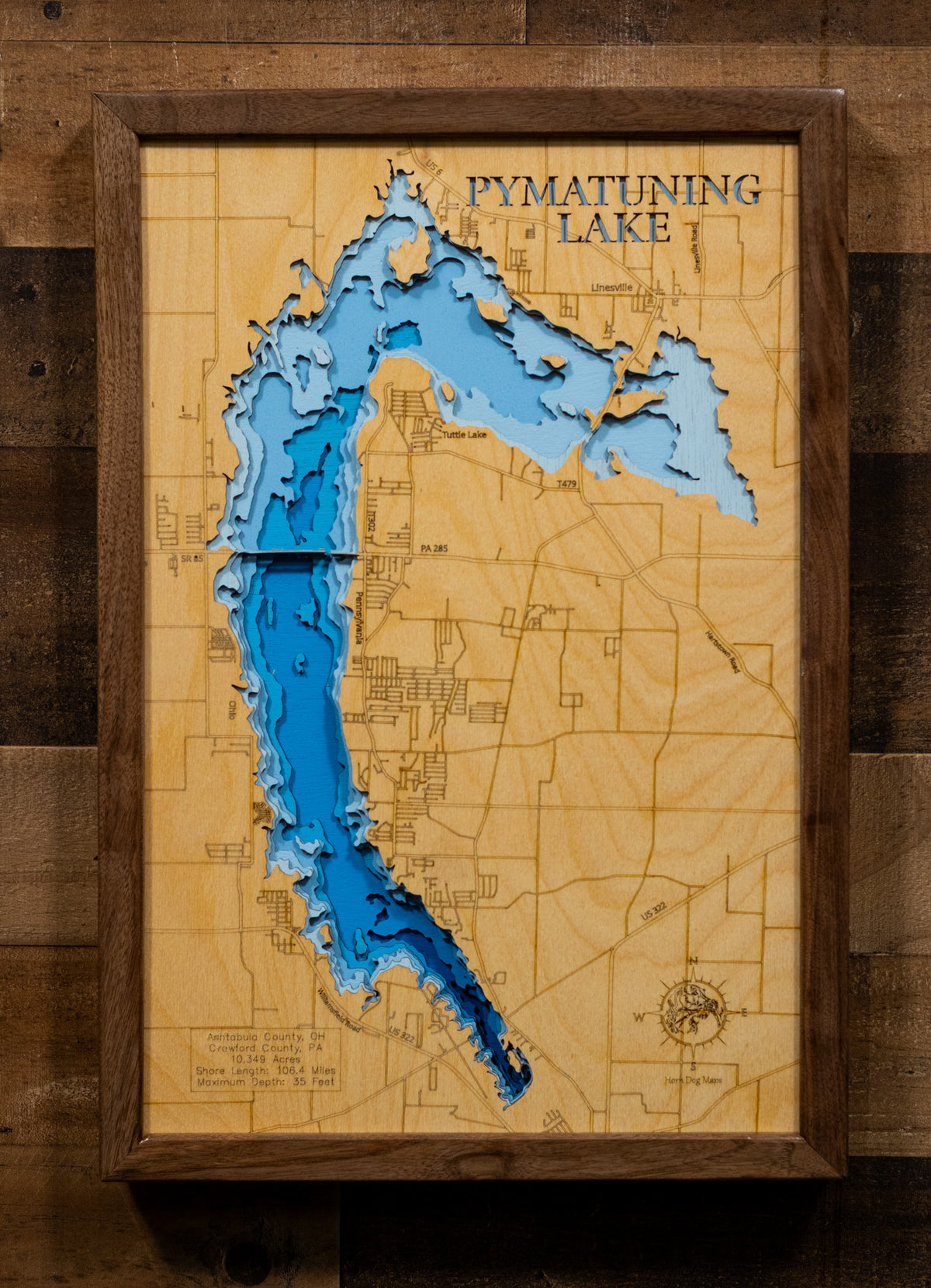 A framed 3D laser-cut wooden map of Pymatuning Lake, featuring layered blue and light blue wood sections over a natural wood background, mounted on a dark wood plank wall.