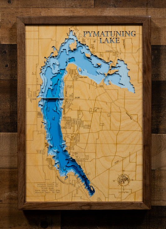 A framed 3D laser-cut wooden map of Pymatuning Lake, featuring layered blue and light blue wood sections over a natural wood background, mounted on a dark wood plank wall.