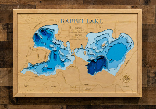 A 3D wooden topographic map of Rabbit Lake, featuring layered blue wood sections representing different water depths on a natural wood background, mounted on a rustic dark wood plank wall.