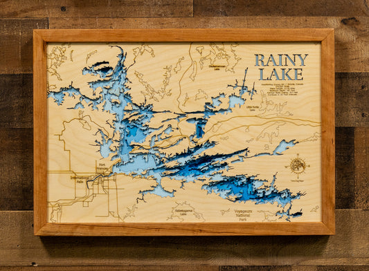 A framed wooden depth map of Rainy Lake, featuring a light natural wood background, blue laser-engraved water channels, and a light wood frame, mounted on a dark rustic wood plank wall.