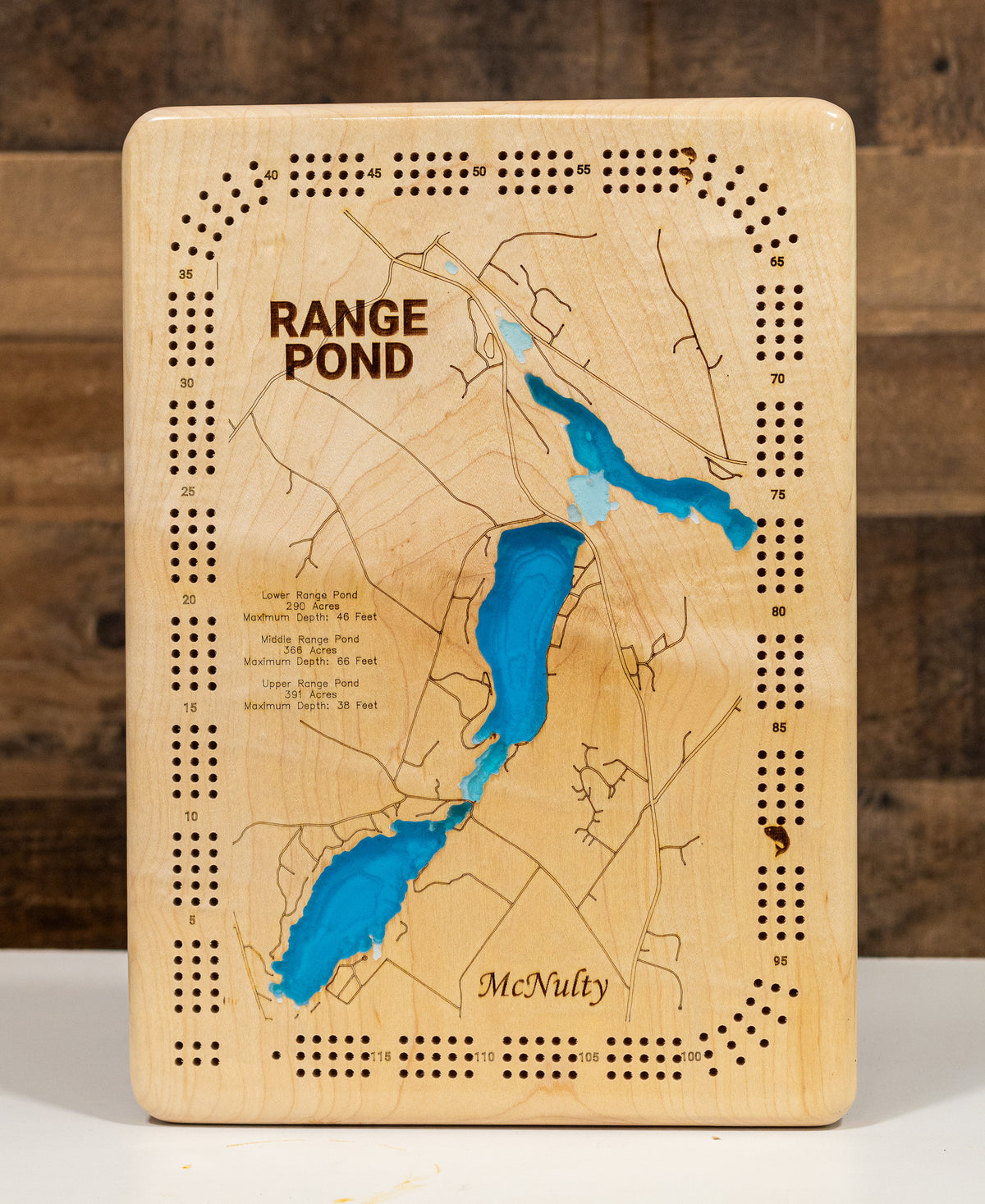 A cribbage board game featuring an engraved map of Range Pond with blue water channels, laser-cut holes around the border, and the names 'Range Pond' and 'McNulty' printed on the front. The board is photographed upright against a rustic dark wood background.