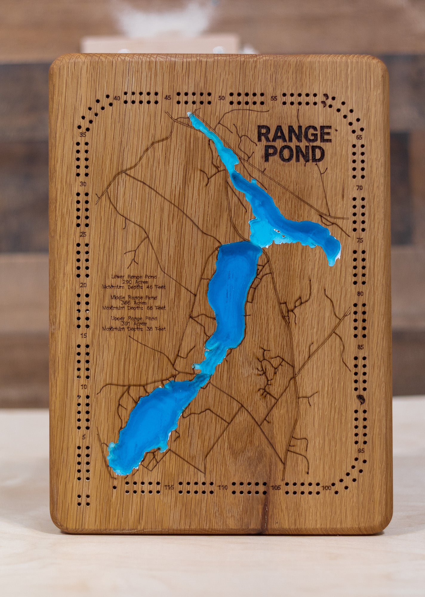 A highly detailed, laser-cut, and multi-layered wooden map of Upper, Middle, and Lower Range Ponds, an interconnected three-lake chain in Poland, Maine. The map shows the distinct shapes of the ponds and their extensive combined shoreline, highlighting the location of Range Ponds State Park on Lower Range Pond. The varying depths of the ponds, reaching a maximum of 66 feet in Middle Range Pond, are represented by different layers of wood.