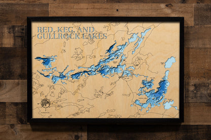 A framed 3D laser-cut wooden map of Red, Keg, and Gullrock Lakes, mounted on a rustic dark wood plank wall. The map features natural light wood grain with layered blue water depths and dark blue text at the top.