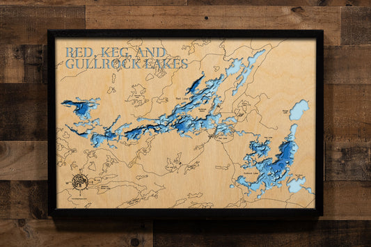 A framed 3D laser-cut wooden map of Red, Keg, and Gullrock Lakes, mounted on a rustic dark wood plank wall. The map features natural light wood grain with layered blue water depths and dark blue text at the top.