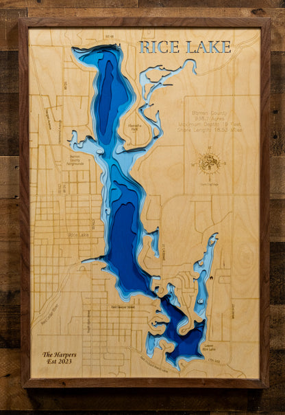 A framed wooden depth map of Rice Lake, featuring layered blue and dark blue resin over a light natural wood background, mounted on a rustic dark wood wall.